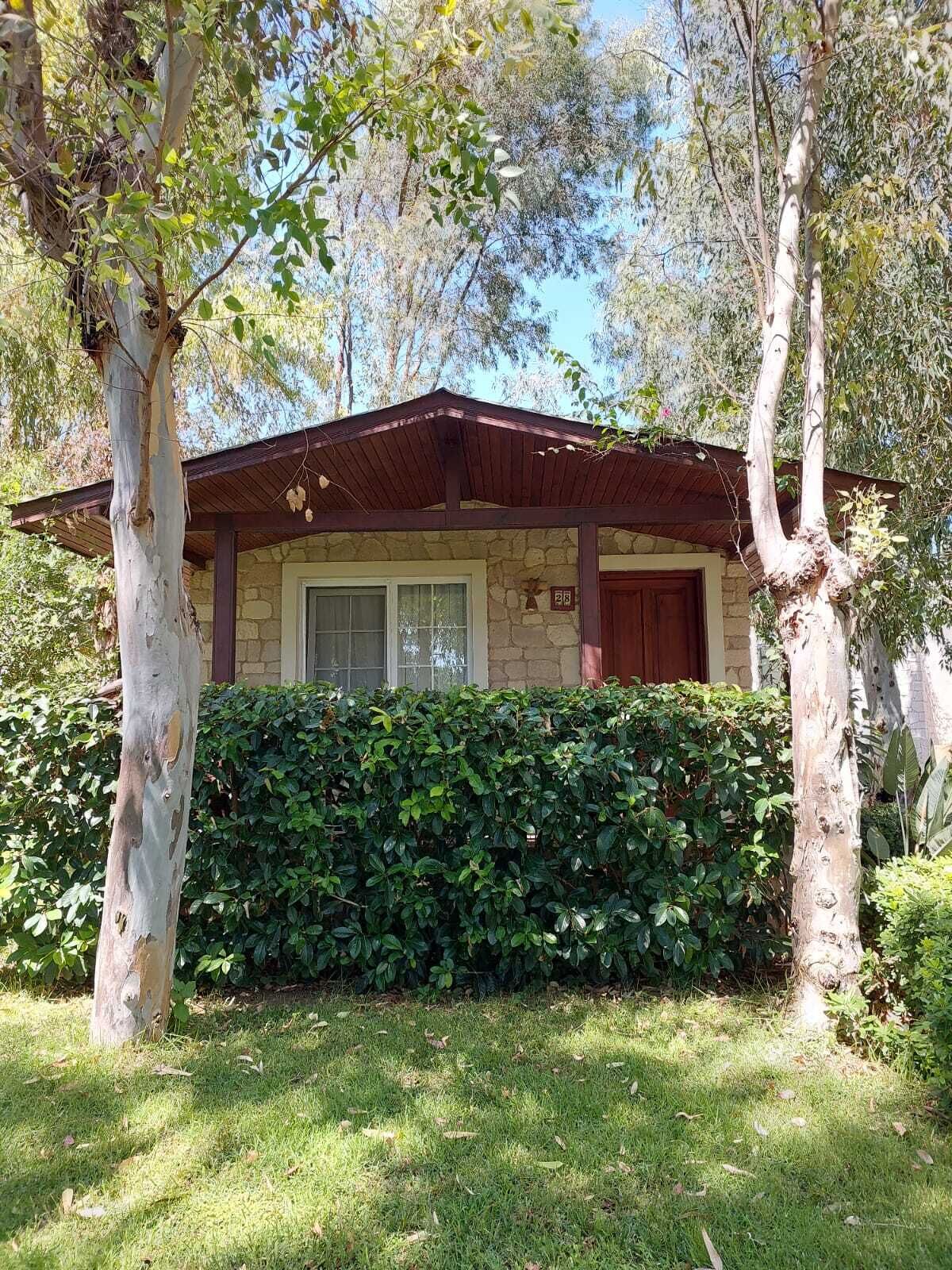 A small stone house with a wooden roof, surrounded by lush greenery and trees.