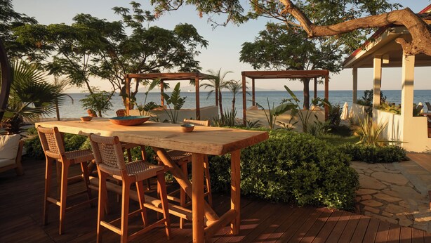 A wooden deck with a table and chairs, surrounded by greenery and overlooking a body of water.