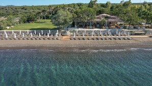 An aerial view of a coastal resort with clear waters, a beach with umbrellas, and a building with a tiled roof.
