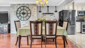 Un coin repas avec une table dressée pour six personnes, une grande horloge murale et, au fond, une cuisine moderne.