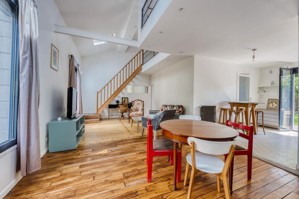 A spacious room with a wooden floor, a dining table with chairs, a staircase, and a large window.