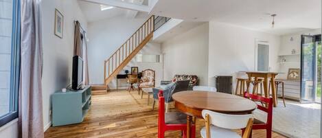 A spacious room with a wooden floor, a dining table with chairs, a staircase, and a large window.