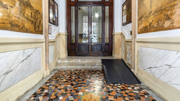 Un pasillo con suelo de baldosas, una puerta de madera con paneles de vidrio y dos cuadros en las paredes.