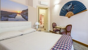 Habitación de hotel con cama, escritorio con silla, ventilador y un cuadro de un atardecer sobre un río.