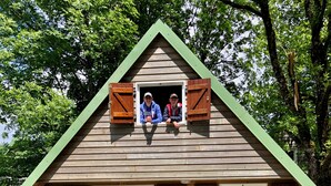 Een houten chalet met een puntig dak, een voordeur en een klein terras met plastic stoelen.