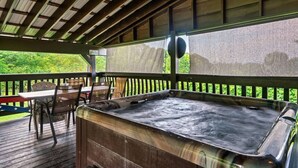 Ein Whirlpool auf einer Holzterrasse mit Tisch und Stühlen, umgeben von einem Hollerleger und einem überdachten Bereich mit Blick auf Grünflächen.