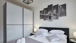 Moderni hotellihuone, jossa on suuri sänky, harmaat vuodevaatteet, valkoiset tyynyt ja seinällä kaupunkimaisemaa esittävä taideteos.