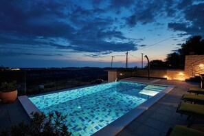Ein Schwimmbecken mit geometrischem Fliesenmuster, umgeben von Liegestühlen und Topfpflanzen.