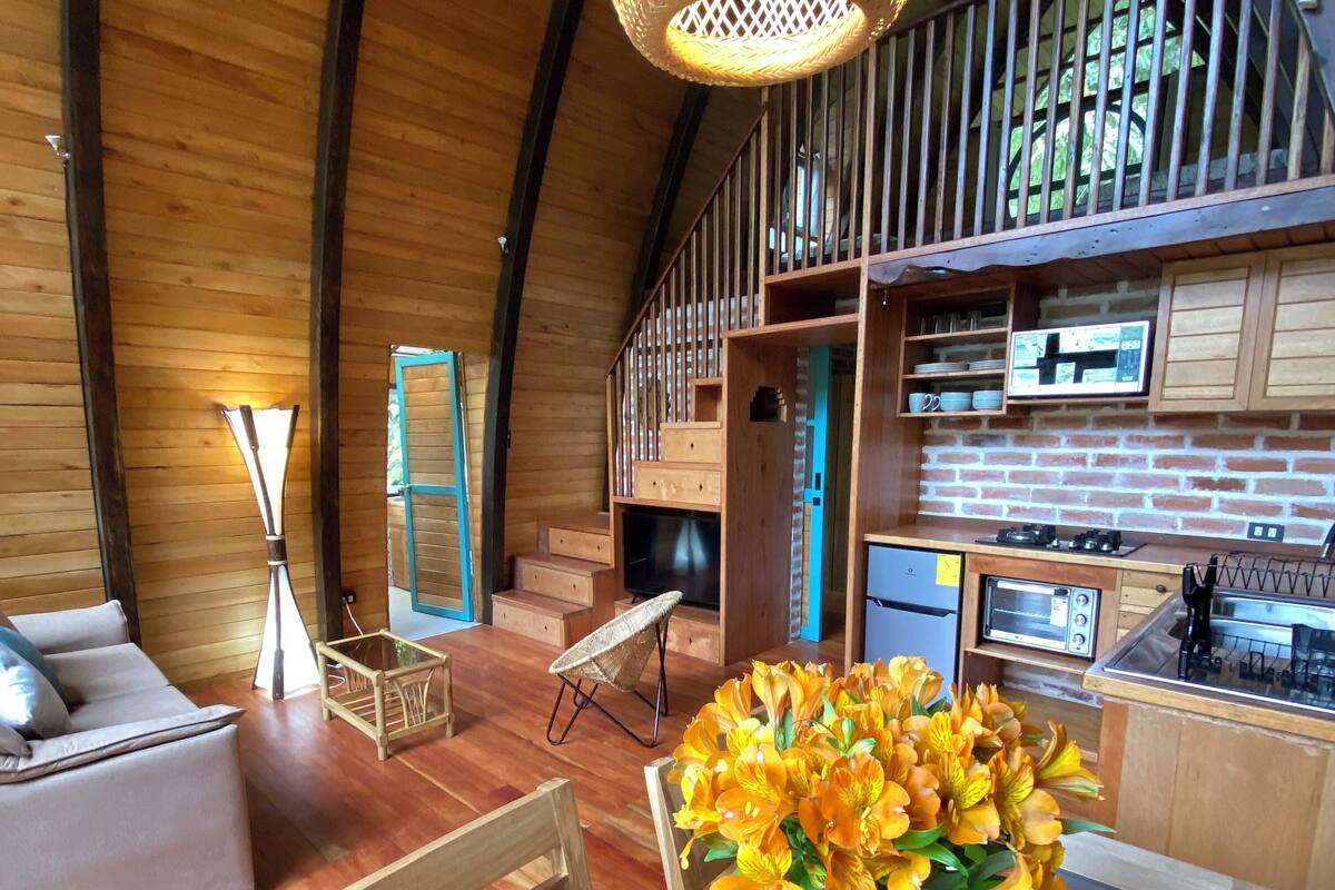 Interior acogedor de una cabaña con comedor, muebles de madera, lámpara colgante y cocina con salpicadero de ladrillo.