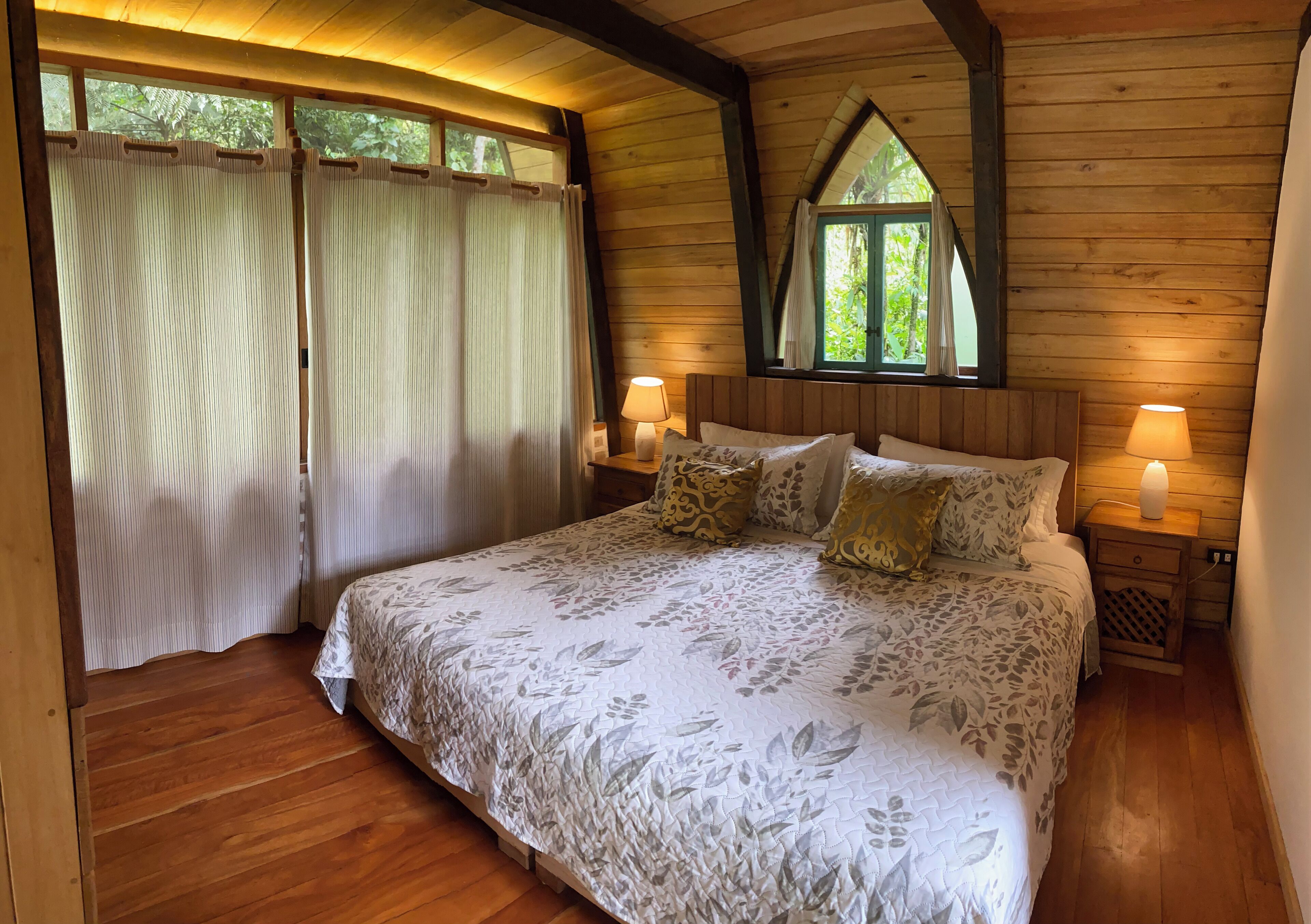 Quarto com cama grande, duas mesas de cabeceira com luminárias, janela com vista para o verde e paredes de madeira.