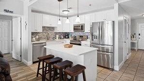 A modern kitchen with white cabinetry, stainless steel appliances, and a central island with bar stools. - Ocean Views-5 Pools-Hot Tub-Shops LTW 1006 (Panama City Beach)