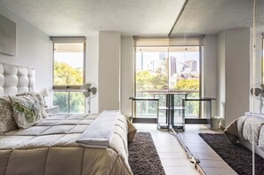 A modern bedroom with a large bed, a small table, and a view of the city through floor-to-ceiling windows.