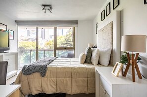 Enjoy a city view from the large window in this stylish bedroom, featuring a comfortable bed, a desk for work, and a touch of greenery.