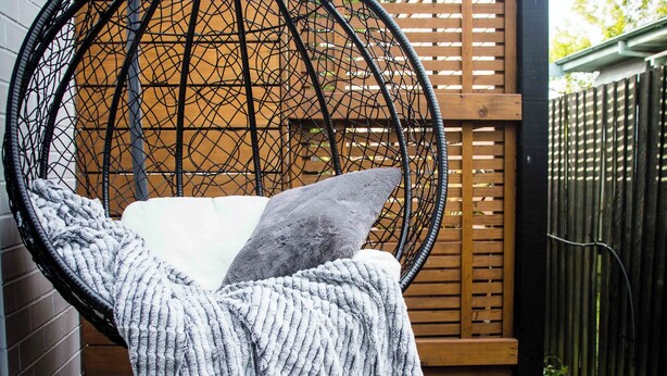 Ein Hängestuhl mit Kissen und Decke, eine Holzterrasse und ein Holzzaun im Hintergrund.