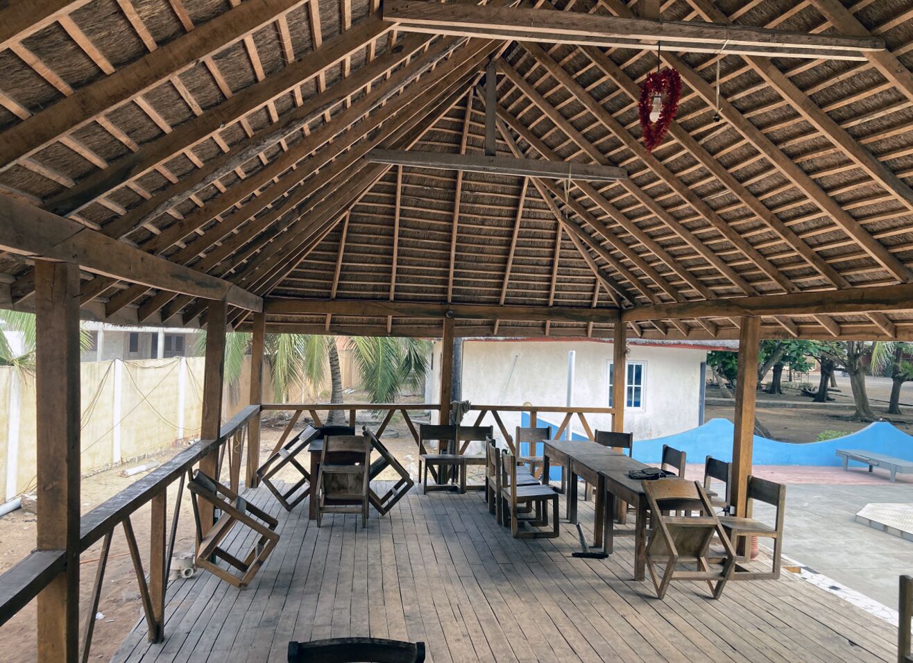 Un espacio al aire libre techado con mesas y sillas de madera, techo de paja y vista de un edificio al fondo.