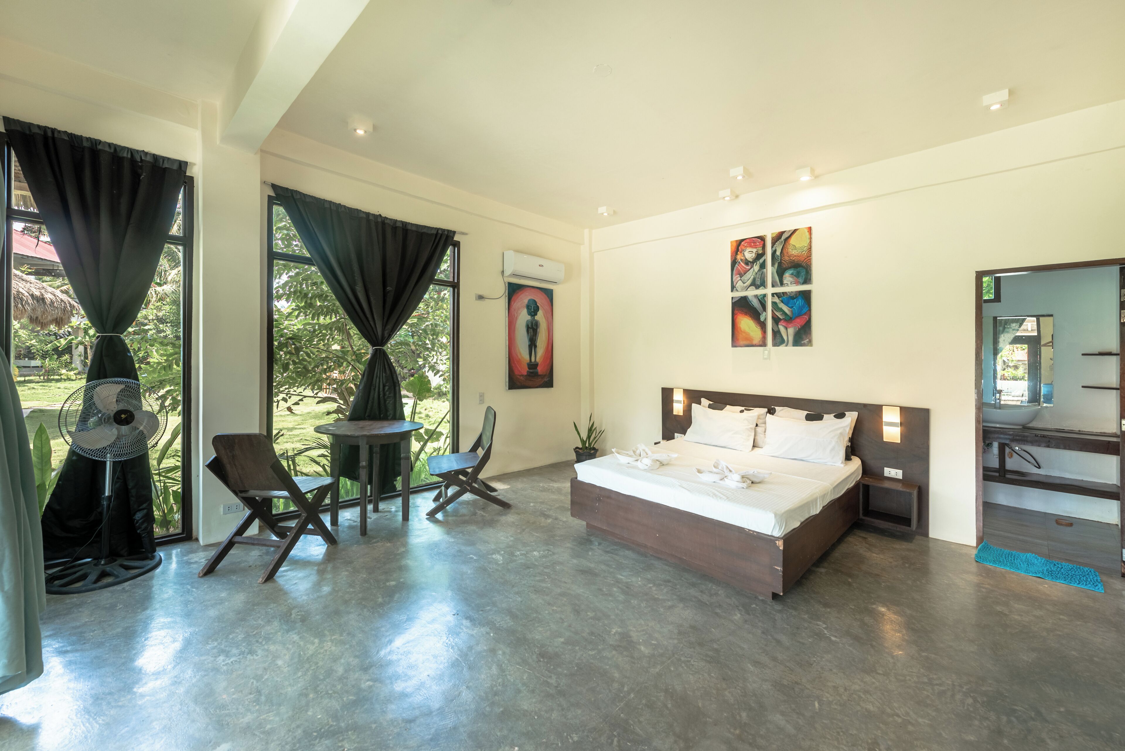 A modern hotel room with a large bed, a small table, two chairs, a fan, and a view of the outdoors.
