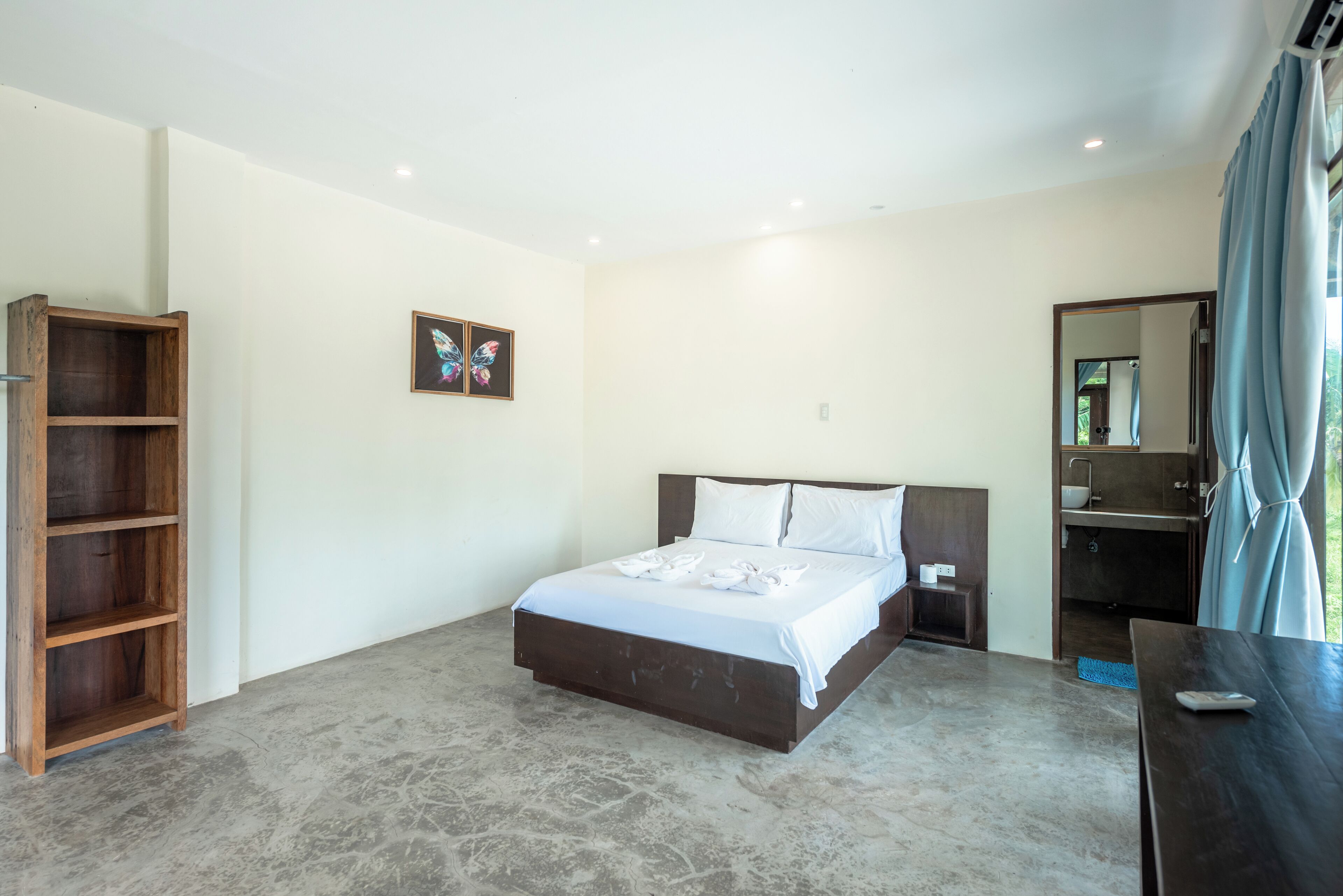 A bedroom with a bed, a wooden shelf, a small bedside table, a mirror, and a bathroom visible through a door.