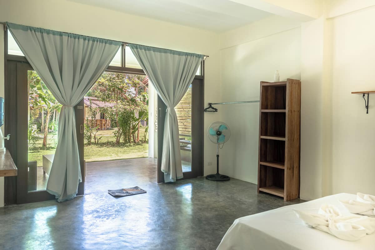 A room with a bed, a wooden shelf, a fan, and a view of the outdoors.