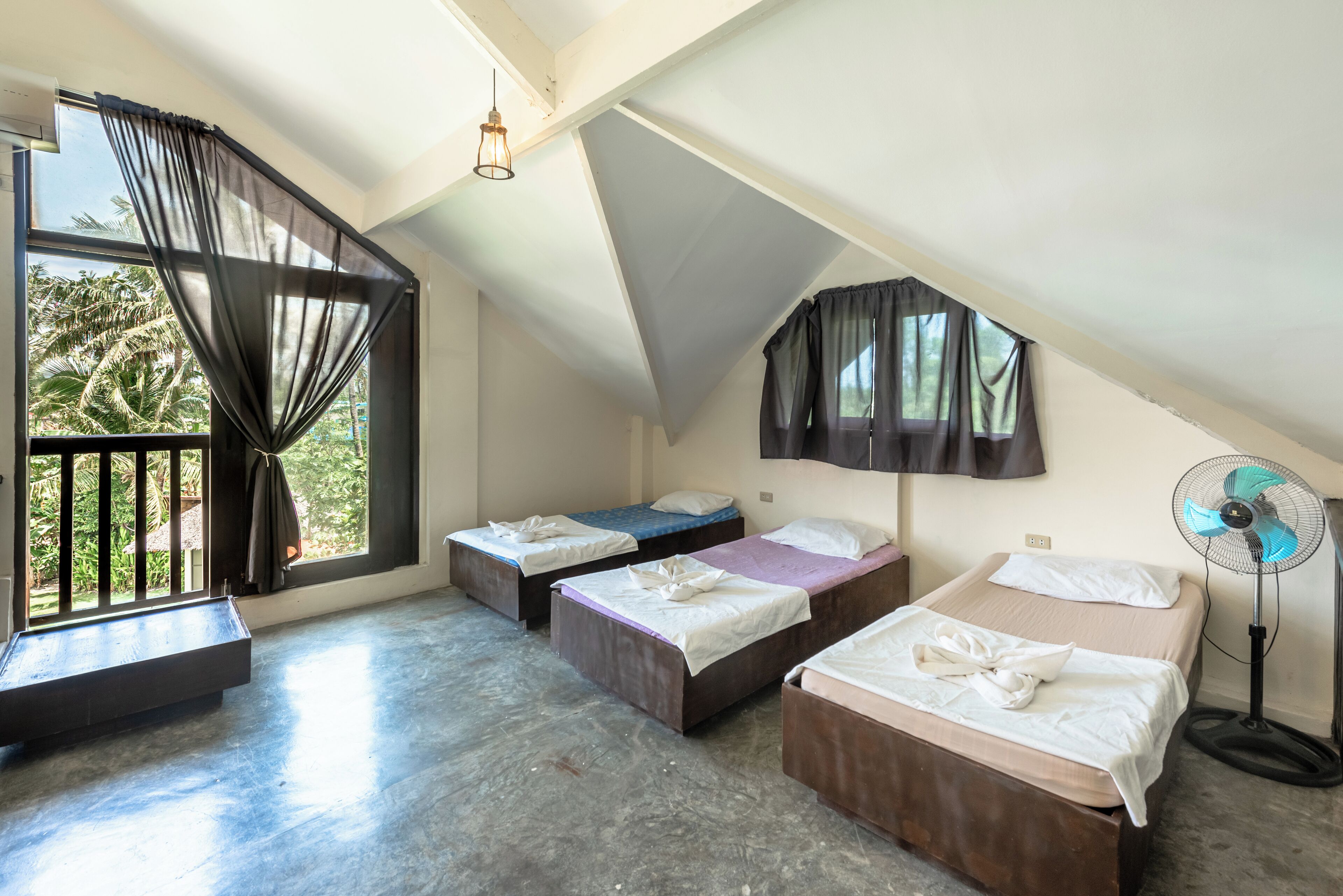 A dormitory room with three beds, a standing fan, and a large window with curtains.