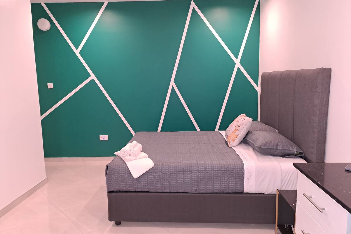 A modern bedroom with a geometric wall design, a grey bed with white bedding, and a dark headboard.