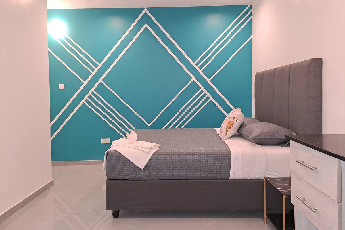 A modern bedroom with a geometric wall design, a grey bed with a white blanket, and a grey headboard.