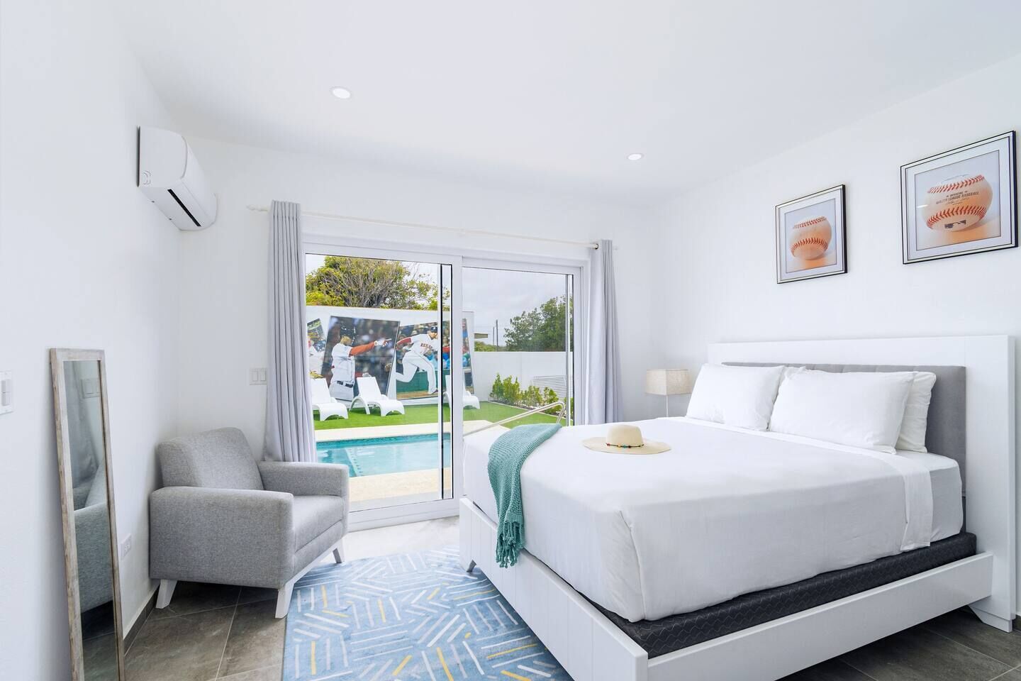 Ein modernes Hotelzimmer mit einem großen Bett, einem grauen Sessel, Blick auf einen Pool und Grünflächen sowie gerahmten Sportmemorabilien an der Wand.