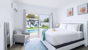 A modern hotel room with a large bed, a grey armchair, a view of a pool and greenery outside, and framed sports memorabilia on the wall.