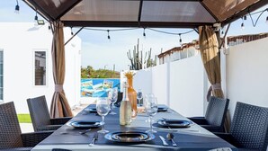 An outdoor dining area with a covered table set for four, featuring wicker chairs, a bottle of olive oil, and wine glasses.