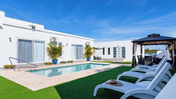 A modern outdoor pool area with a clear blue sky, a pool, white lounge chairs, and a covered seating area.