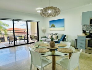 A modern living room with a glass dining table, white chairs, a sofa, and a kitchen area with cabinets and a stove.
