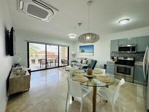 A modern kitchen with stainless steel appliances, a dining area with a glass table, and a living room with a sofa and a view to the outside.