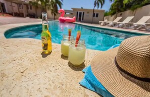 A poolside setting with a bottle, two glasses of a yellow beverage, a straw, and a straw hat on a textured surface.