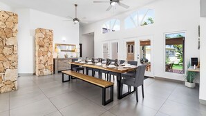Een ruime eetruimte met een lange houten tafel, zwarte stoelen en een bank. Twee plafondventilatoren, stenen muren en grote ramen met uitzicht op de omgeving.