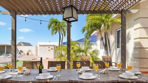 Een overdekt terras met eettafels gedekt voor het ontbijt, met zicht op palmbomen en gebouwen op de achtergrond.