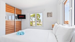 Een hotelkamer met een bed, een aan de muur bevestigde televisie, een raam met uitzicht op palmbomen en een houten garderobekast.