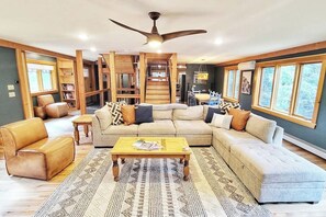 A spacious living room with a large sectional sofa, a coffee table, and a ceiling fan. There are bookshelves and a dining area visible in the background. - Stunning New Build on the River, 4 miles to Trapp (Stowe)