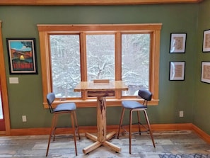 A dining area with a wooden table and two chairs, a large window with a view of trees, and framed artwork on the walls.