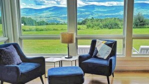 Ein Zimmer mit zwei Sesseln und einem kleinen Tisch, große Fenster mit Blick auf grüne Felder und Berge.