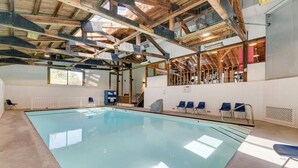 An indoor swimming pool with a high ceiling and visible wooden beams, surrounded by blue chairs and a white wall.