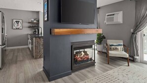 A modern living room with a fireplace, a mounted TV, and a kitchen in the background.