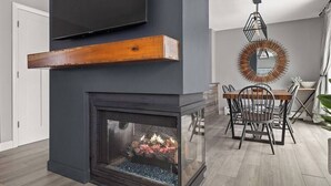 A modern living room with a fireplace, a dining area with a wooden table and chairs, and a large mirror on the wall.