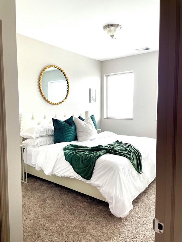 A neatly made bed with white bedding and a green throw blanket, a round mirror with a gold frame, and a window with blinds.