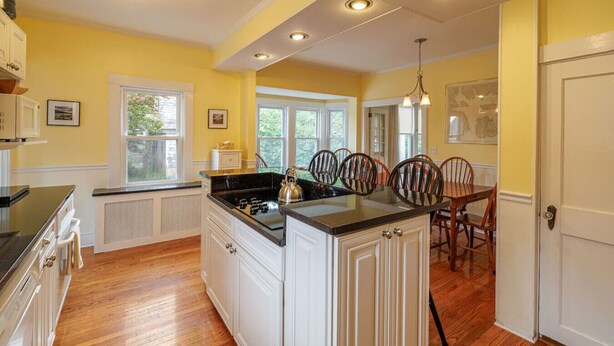 A kitchen with white cabinetry, a central island, and a dining area with wooden chairs. - Family Pet Friendly 4BR in N.E. Harbor[Millbrook] (Mount Desert)