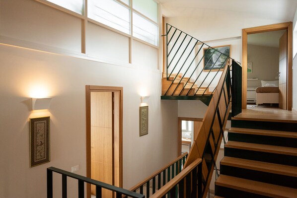 A staircase with wooden steps and black railings, leading to an upper floor. There are wall-mounted light fixtures and framed pictures on the walls.