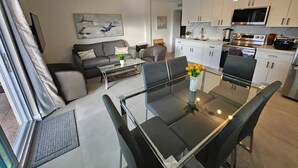 Un appartement moderne comprenant un salon, un coin repas et une cuisine. On y trouve un canapé, une table basse et une table à manger en verre avec des chaises. La cuisine est équipée de meubles blancs, d’une cuisinière et d’un four à micro-ondes.