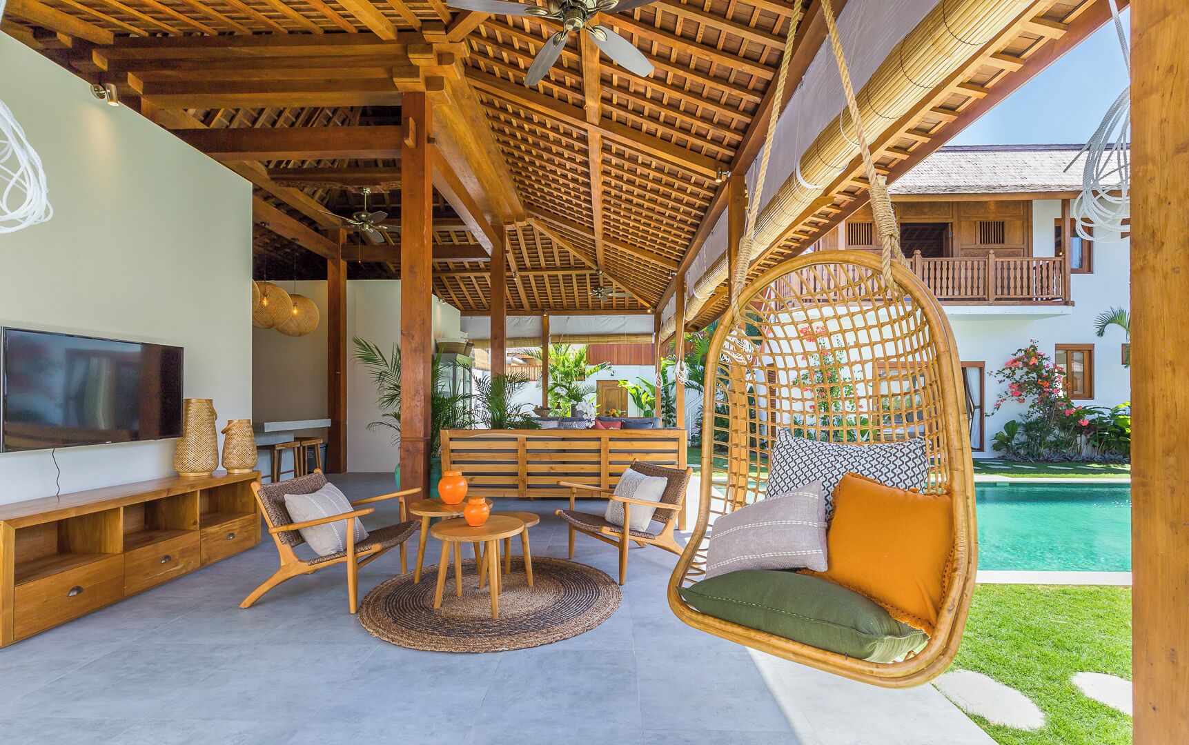 A cozy outdoor seating area with wicker chairs and a hanging chair, a TV, and a pool in the background.