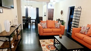 Un salón con un sofá naranja, una mesa de centro y un televisor en la pared. Se ve un ventilador de techo y una puerta que da a otra habitación.