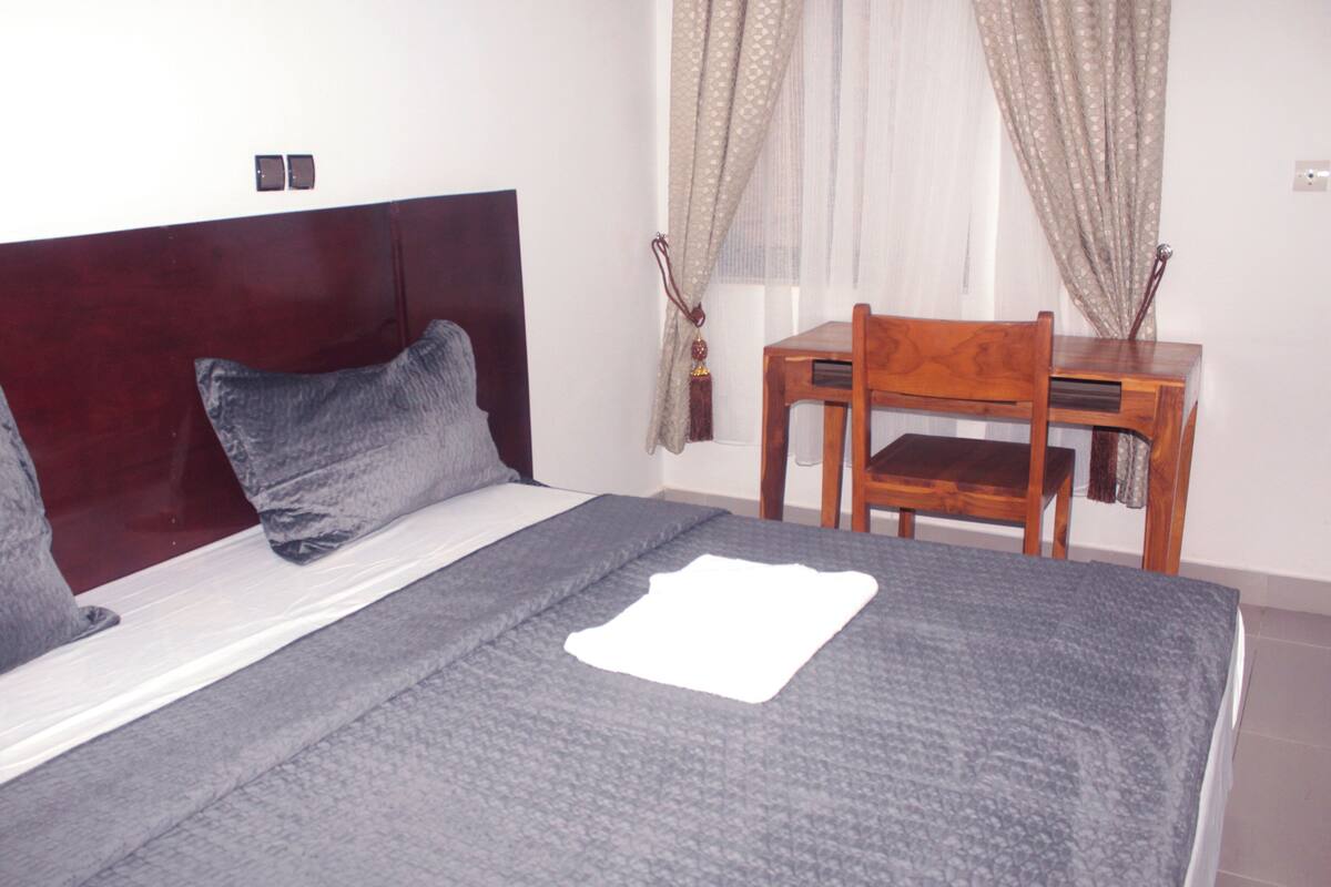 A bed with a wooden headboard, two pillows, and a blanket. There is a wooden chair and a small table beside the bed.