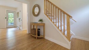 A hallway with wooden flooring, a staircase with a wooden handrail, a round mirror on the wall, and a small table with a plant and baskets. - Quiet Family & Pet Friendly Home! [Owls Nest] (Tremont)