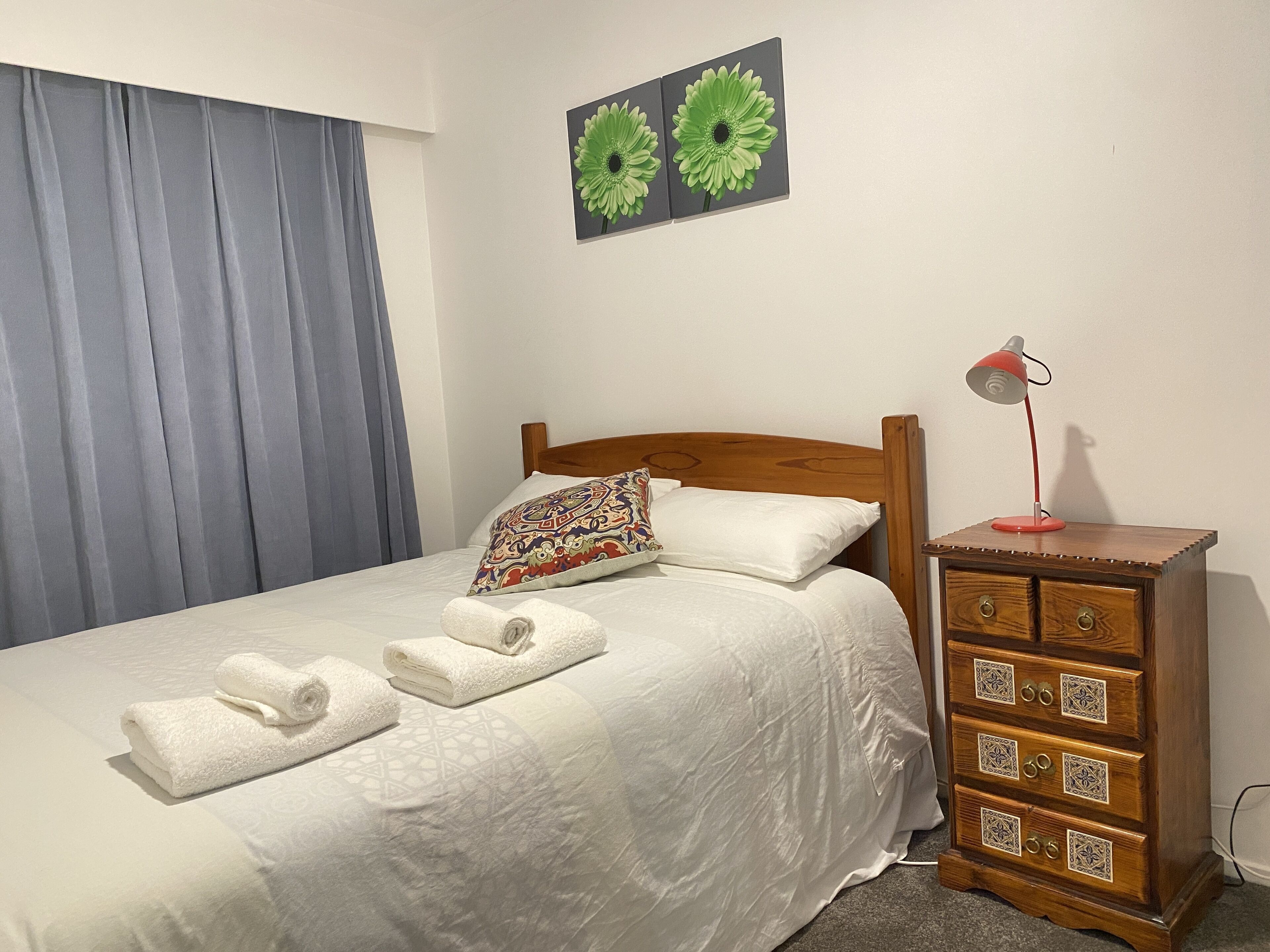 Un lit bien fait, avec du linge de lit blanc et deux serviettes enroulées, une tête de lit en bois, un coussin à motifs, une petite commode en bois avec des tiroirs et une lampe de bureau rouge.